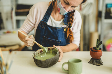 Caucasian woman works with ceramic pottery and color paint while wearing protective face mask for coronavirus outbreak