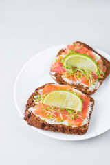 Two toast sandwiches on dark grain bread with butter, lightly salted salmon, microgreens. Idea for healthy breakfast, snack. Served on white plate, light background with lime. Healthy fats omega 3.