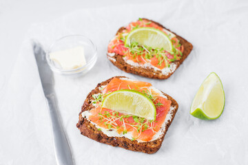 Two toast sandwiches on dark grain bread with butter, lightly salted salmon, microgreens. Idea for healthy breakfast, snack. Healthy fats, diet ketogenic recipe. Served on light background with lime.