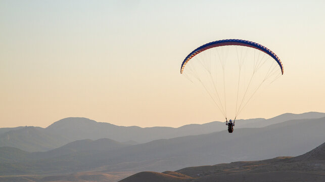 AERIAL: A man on a paraglider flies during sunset over a valley. The last rays of the sun illuminate his wing. A sense of freedom fills the chest.