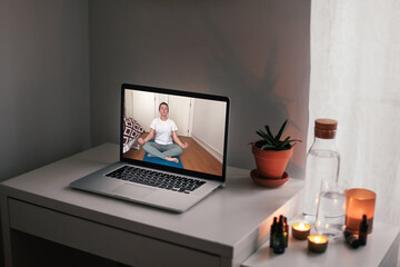 Woman meditating online at home. A video with remote yoga in a laptop's screen. A laptop with a video call on a table at home, candid atmosphere