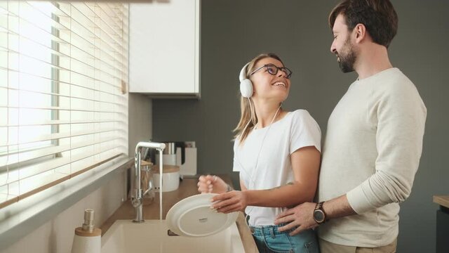 A smiling man is hugging his girlfriend while she is washing the dishes at home