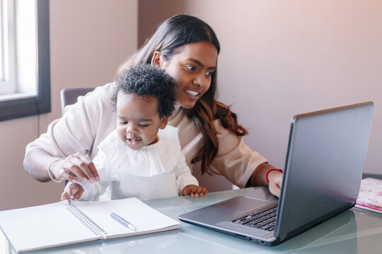 Mixed race Indian mother with African black baby working online from home on Internet. Workplace of freelancer woman with kid. Stay home single mom working distant job. A new normal.