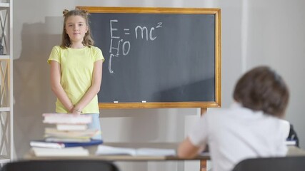 Confident intelligent Caucasian schoolgirl writing physical formulas on chalkboard in classroom and turning to camera. Blurred schoolboy sitting at desk at front. Children studying in school. - Powered by Adobe
