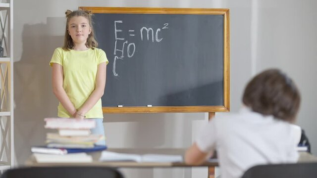 Confident intelligent Caucasian schoolgirl writing physical formulas on chalkboard in classroom and turning to camera. Blurred schoolboy sitting at desk at front. Children studying in school. - Powered by Adobe