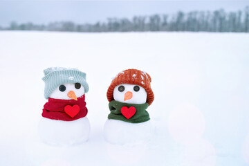 Valentines day card with a snowmans. Two little cute snowman in a orange hat and a green scarf with red hearts in the snow, close-up. 