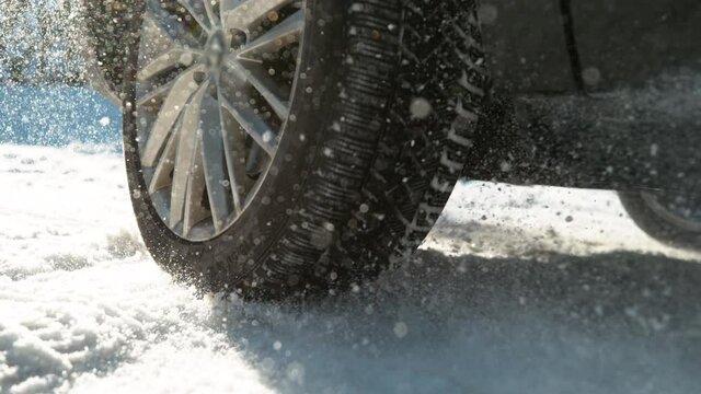 SLOW MOTION, LOW ANGLE, CLOSE UP, DOF: Fresh snow flies up from a large vehicle's spinning wheel. Car's wheels spin and spew up pieces of snow as it attempts to gain traction on the slippery road.