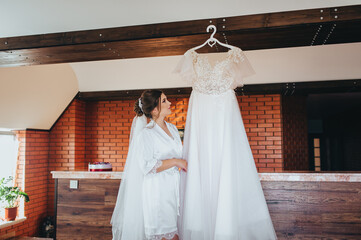 A beautiful bride in a white robe stands in the interior and looks at a wedding dress hanging on a...