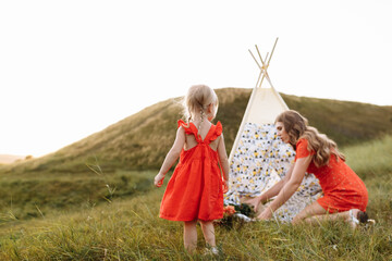 Beautiful mother with her little daughter sitting near wigwam in the field. Spending time together, outside, on vacation, outdoors. Beautiful sunset light in the garden or in the park © Andriy Medvediuk