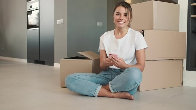 A smiling woman is using her smartphone while sitting on the floor near many boxes at home