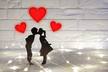 black silhouette kissing a couple of guy and girl, above them three red hearts, around the pair glows garland, in the background a white brick wall