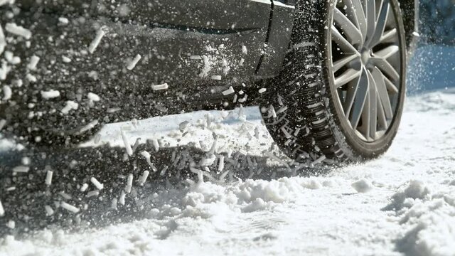 SLOW MOTION TIME WARP, LOW ANGLE, CLOSE UP, DOF: Car's wheels spin in place while trying to drive along snowy street. Vehicle struggling to gain traction on the winter road spews up pieces of snow.