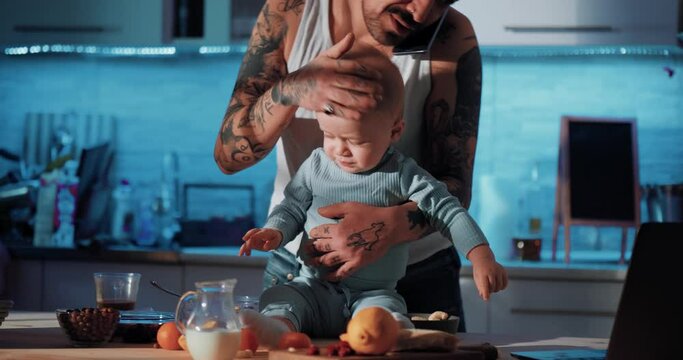 Happy baby on the table laughing, young dangerous dad taking care of the child. A busy parent talks on the phone and takes care of the baby. Dad and baby in the kitchen