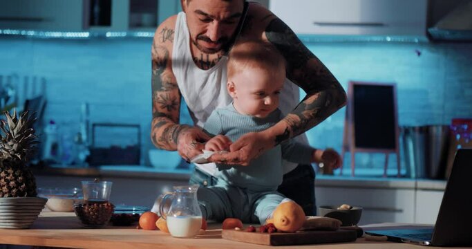 Tattooed dad wipes the baby's hands and talks on the phone. Dad and baby in the kitchen, preparing food and using laptops. Baby on the table