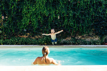 Little boy jumping into a pool. His mom looking hi. Child get fun in the swimming pool of his home....