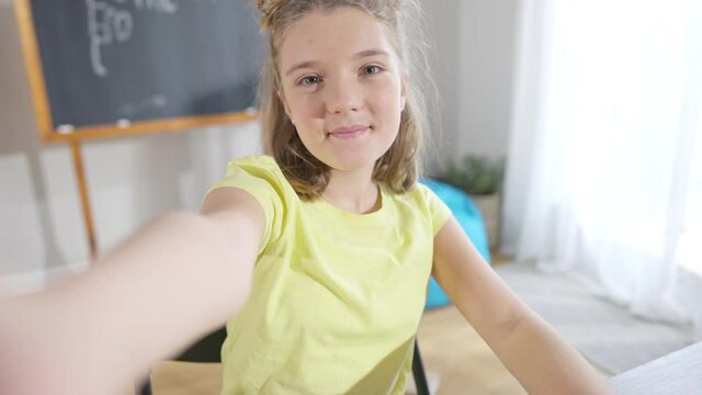 Cheerful beautiful Caucasian girl taking selfie in classroom looking at camera and showing peace gesture. Portrait of charming positive schoolgirl having fun on break in school. Generation Z.