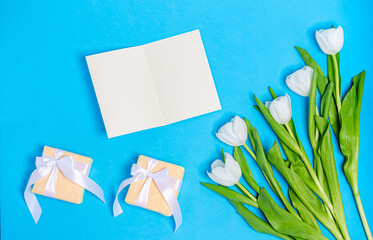 White tulips bouquet with greeting card and gift boxes on a blue background.