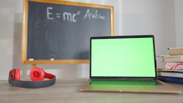 Close-up of laptop with chromakey screen on desk in classroom with headphones and blurred chalkboard at background. Green screen device indoors at school. Modern technologies concept.