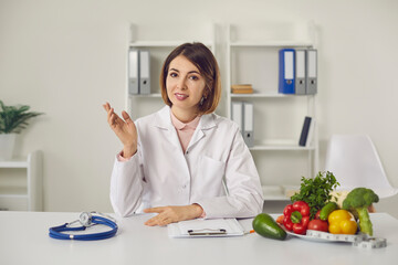 Dietitian, nutritionist or doctor sitting at desk at the clinic, looking at camera and telling about benefits of eating fresh fruit and vegetables, sharing weight loss advice and healthy diet habits