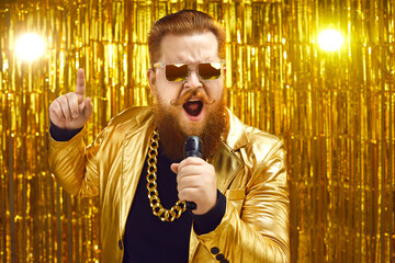 Funny singer in shiny golden jacket and gold chain holding microphone and singing songs at retro pop music concert. Happy bearded man performing at disco nightclub or enjoying time at karaoke party - obrazy, fototapety, plakaty