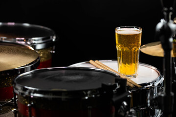 Glass of light beer on professional drum set closeup. Drumsticks, drums and cymbals, at live music rock concert, in the club stage, bar, or in recording studio. Black background.