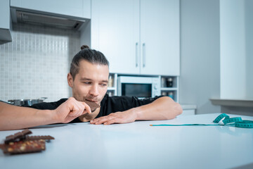 sportive man on diet with chocolate bar and measuring tape at modern kitchen