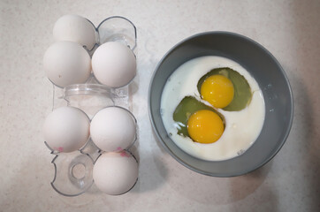 Two raw eggs in a bowl with milk near white eggs in eggshell in egg tray. Food ingredients for cooking omelette for breakfast
