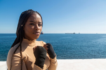 Portrait of beautiful young afro american woman looking at camera, with bandages on her hands to practice martial arts, boxing. Sky and sea background, seafront.