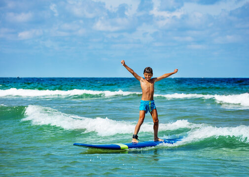 Young 10 years old boy posing with lifted hands ride surfboard on a wave in the sea