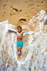 Sea waves touch happy relaxed boy lay on the sand with stretched hands wearing sunglasses view from above