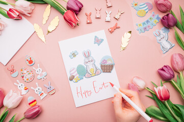 Hand is writing Happy Easter on card. DIY ideas and step by step instructions for making Easter Card. How to make handmade card for beginners with stickers and flowers nearby