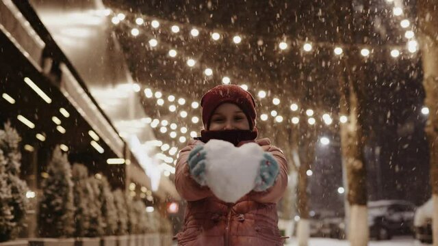 Portrait of a young enthusiastic girl on the Christmas decorated street during a snowfall with a snow heart in her hands