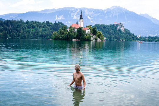 Latin skinny boy with dyed white hair in sunglasses in swimwear in a blue lake and a island with a church behind