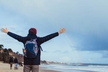 Fototapeta na wymiar Back View man in warm clothes raised arms up to the sky, enjoying rainbow view above the sea during a walk on winter seaside. Unity with nature, digital detox. Local self-traveling in autumn, winter.