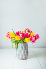 Fresh spring yellow and pink tulips bouquet in blue vase on white wooden table with white background. Festive flowers for mother's, women's day, birthday. Mockup for vertical greeting card. Copy space