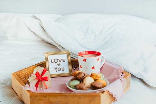 Valentine's day breakfast in bed for Lover. Love you card in the frame, a cup of coffee or cocoa, macaroons, heart-shaped gift box with red ribbon on the wooden tray. Romantic morning. Copy space