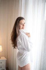 Brunette in mens shirt. Rear view of young woman looking through the window in the morning