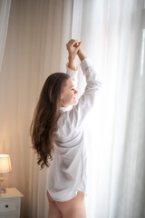 Brunette in mens shirt. Rear view of young woman looking through the window in the morning