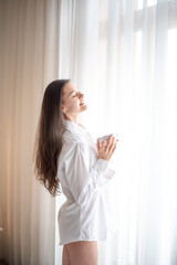 Beautiful brunette woman standing next to a window, wearing men shirt and drinking coffee in the morning