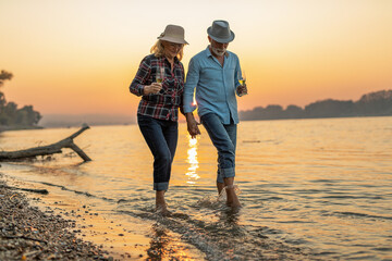 A happy elderly couple walks through shallow water holding hands enjoying the sunset.