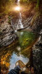 Beautiful waterfall with sunlight in autumn forest. Waterfall vertical panorama. Traveler man stands in front of waterfall river stream.