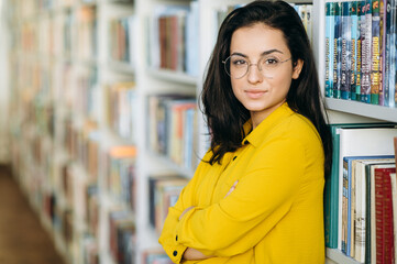 Portrait of elegant young pretty woman. Beautiful female teacher, freelancer or student looks at the camera, smiling. Young stylish lady wearing eyeglasses holds in arms some books, standing in