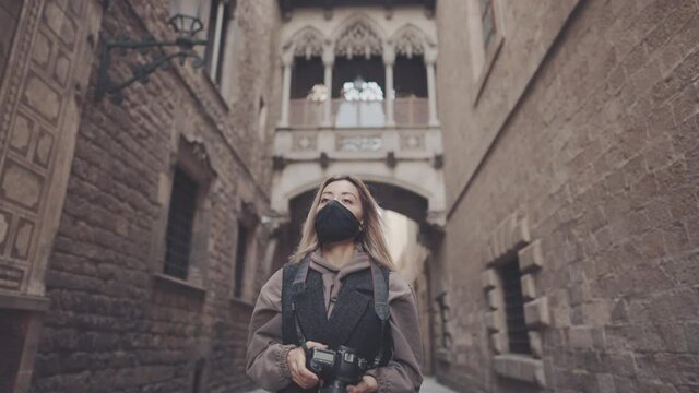 Pandemic portrait of tourist woman wearing mask with dslr photo camera walking through old town narrow streets, Barcelona, Spain. She takes photos. covid tourism concept with coronavirus