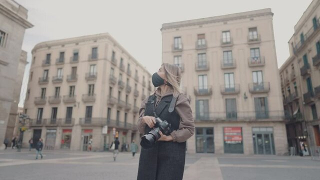 Pandemic portrait of tourist woman wearing mask with dslr photo camera walking through old town square, Barcelona, Spain. She takes photos. covid tourism concept with coronavirus