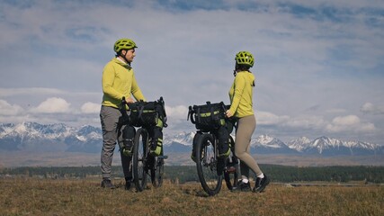 The man and woman travel on mixed terrain cycle touring with bikepacking. The two people journey with bicycle bags. Sport bikepacking, bike, sportswear in green black colors. Mountain snow capped.