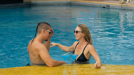 The loving couple hugs and kisses, drinking blue cocktail alcohol liquor in swimming pool at hotel outdoor. Portrait of caucasian man and woman. Creative hairstyles bodybuilder, swimsuit, sunglasses.