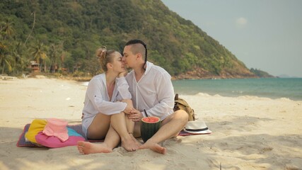 The cheerful love couple holding and eating slices of watermelon on tropical sand beach sea. Romantic lovers two people caucasian spend summer weekend in outdoor. Hat, backpack white shirt beachwear.