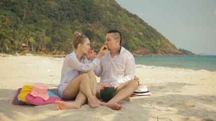 The cheerful love couple holding and eating slices of watermelon on tropical sand beach sea. Romantic lovers two people caucasian spend summer weekend in outdoor. Hat, backpack white shirt beachwear. - obrazy, fototapety, plakaty