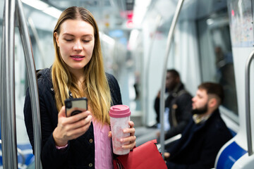 Portrait of positive girl wearing warm coat traveling in metro car and using mobile phone
