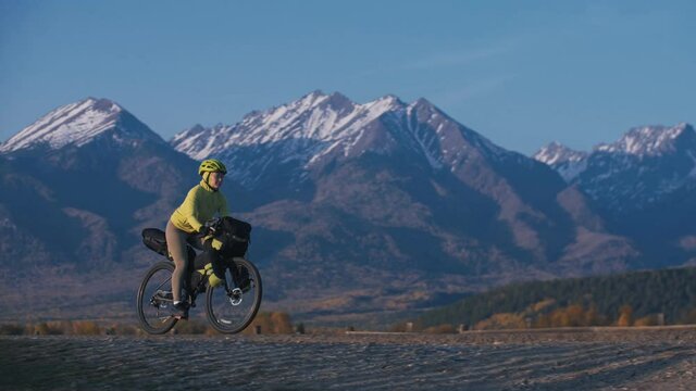 The woman travel on mixed terrain cycle touring with bikepacking. The traveler journey with bicycle bags. Sport tourism bikepacking, bike, sportswear in green black colors. Mountain snow capped.